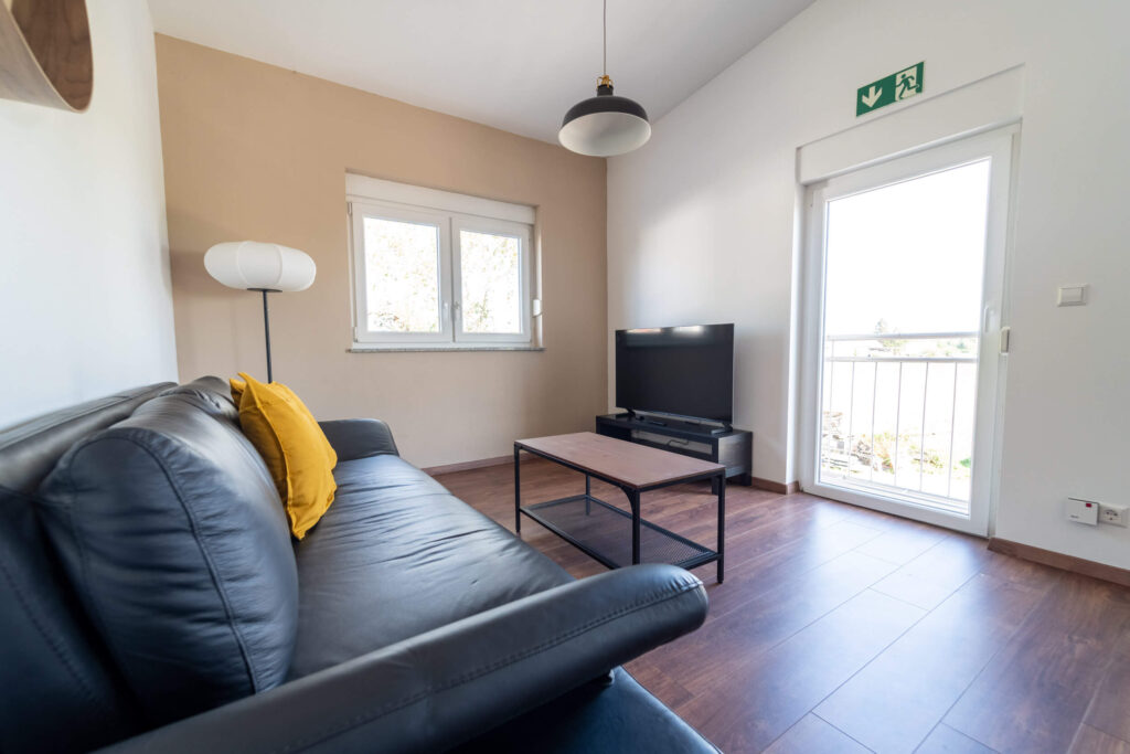 Wohnbereich mit einem schwarzen Sofa, einem Couchtisch und einem Fernsehschrank an der gegenüberliegenden Wand mit Blick aus dem bodentiefen Fenster.